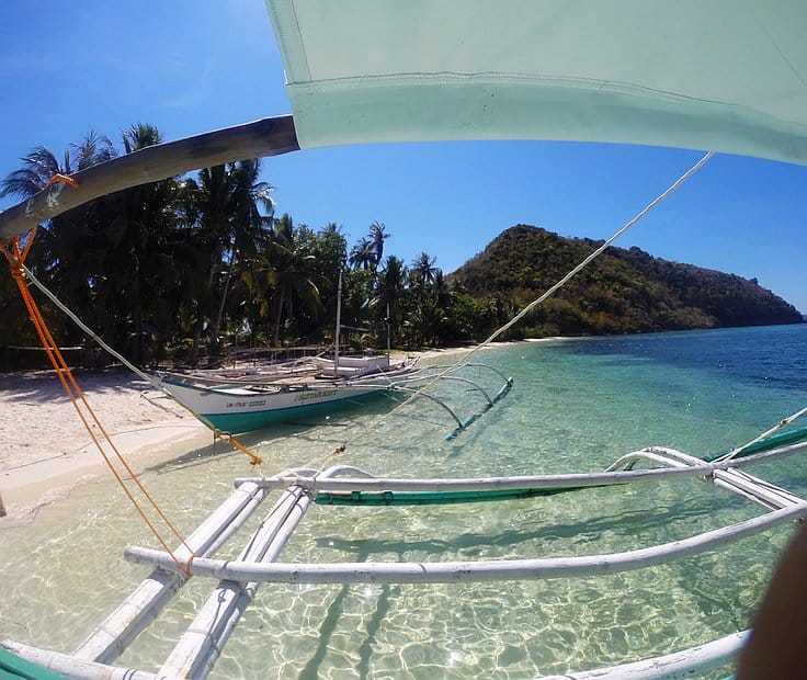 A tranquil beach scene with clear turquoise waters, a small boat anchored near the shore for idyllic island hopping adventures, and lush palm trees lining a sandy beach. A lush, green hill is visible in the background under a clear blue sky.