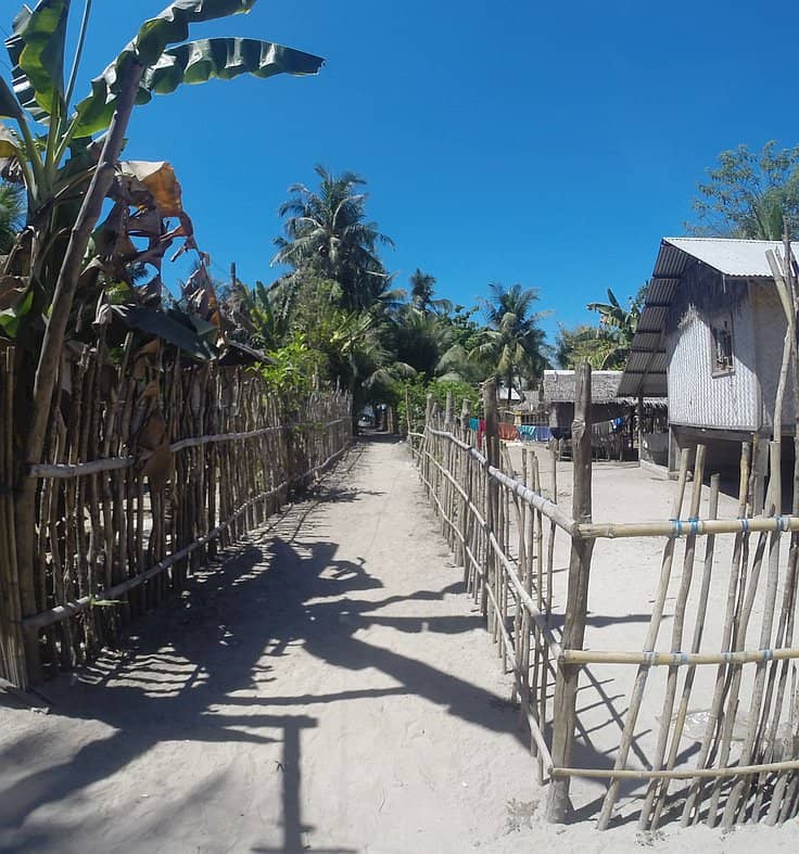 A sandy path lined with bamboo fences leads through a tropical area with lush banana and palm trees. To the right, there's a traditional thatched house on stilts, perfect for resting after a day of island hopping under the clear blue sky.