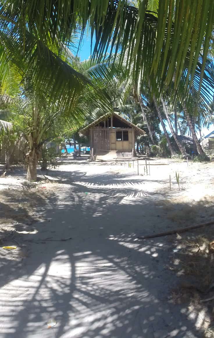 A small wooden hut sits nestled among palm trees on a sandy path. Sunlight filters through the leaves, casting shadows on the ground. Nearby, boat tours depart for island hopping adventures, while the beach and ocean stretch out under a clear blue sky.
