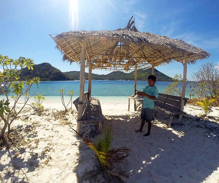 A person stands under a rustic wooden shelter with a thatched roof on a sunny beach, perfect for island hopping. The clear blue sky and calm waters stretch to the horizon, inviting boat tours amidst the green hills in the background. Sparse vegetation surrounds the sandy area.