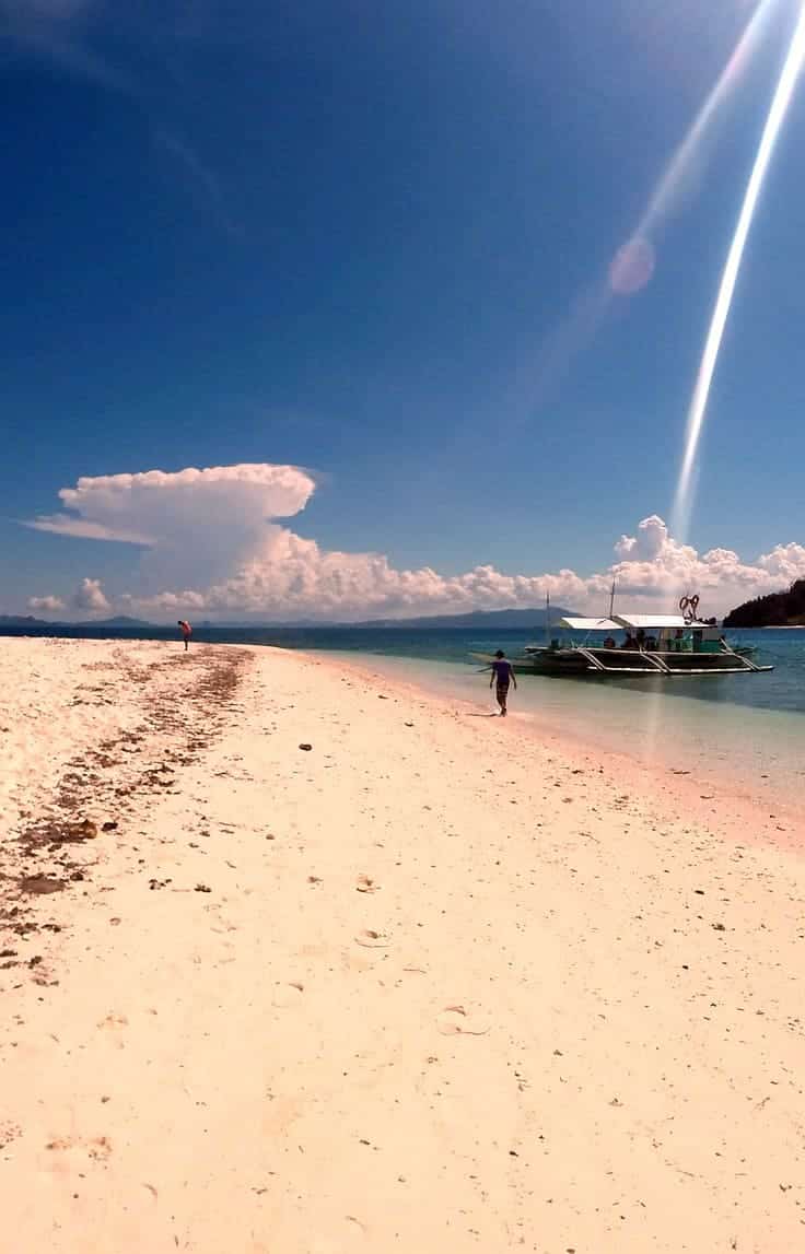 A serene beach scene with white sand, clear blue water, and a partly cloudy sky. A boat anchored near the shore suggests potential for island hopping, while a person walks along the sand. Sunlight creates a bright reflection on the water.