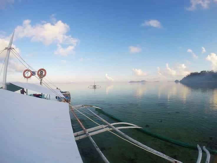 A calm sea under a blue sky with scattered clouds sets the perfect scene for island hopping. A traditional outrigger boat is in the foreground, while another small boat hints at an exciting boat tour in the distance. Islands are partially visible on the horizon.