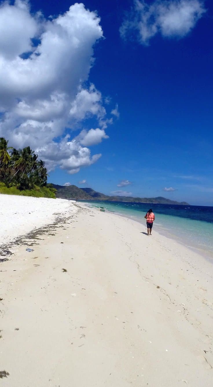 A person walks along a pristine, sandy beach under a vibrant blue sky with scattered clouds. Palm trees line the shore on the left, while boat tours skim the turquoise ocean to the right, meeting distant hills—inviting thoughts of island hopping adventures beyond the horizon.