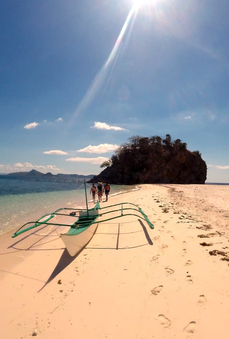 A traditional outrigger boat, perfect for island hopping, rests on a sandy beach under a clear blue sky. People walk near the shoreline, with a small island and distant mountains visible in the background. Sunlight casts shadows across the sand.