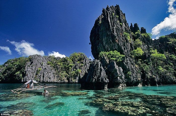 A traditional boat glides on the turquoise waters of El Nido, framed by tall, jagged limestone cliffs under a clear blue sky.