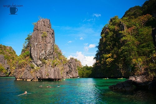 People swimming in clear turquoise waters, surrounded by the tall limestone cliffs and lush greenery of El Nido, under a bright blue sky—a perfect day for a scenic boat tour.