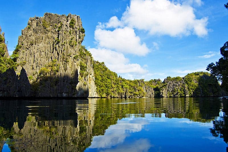 Experience the scenic view of limestone cliffs reflected in tranquil blue water under a bright sky with fluffy clouds during an El Nido boat tour.