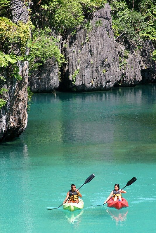Two people kayaking on clear turquoise water surrounded by rocky cliffs and lush greenery, reminiscent of an idyllic boat tour in El Nido.
