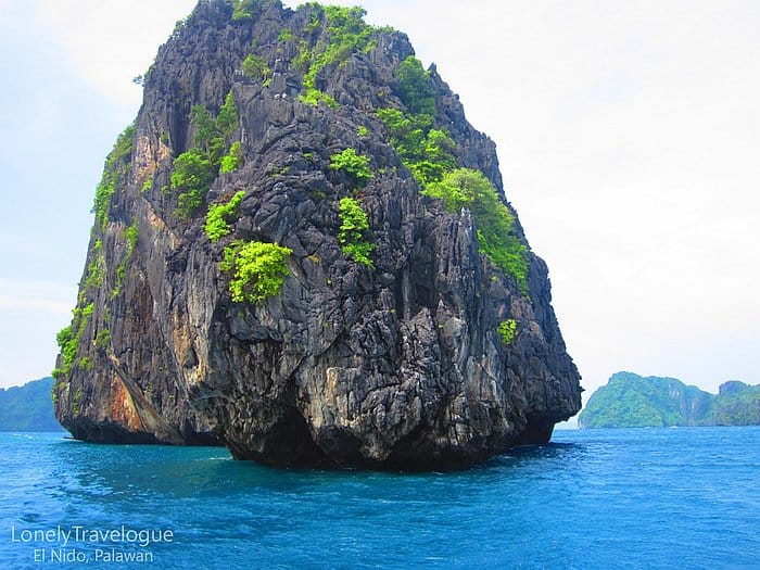 A large, lush rock island rises from the vibrant blue waters under a clear sky in El Nido, where a boat tour offers an unforgettable experience.