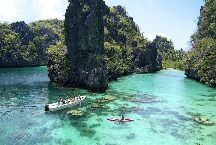 A scenic view of a turquoise lagoon with rock formations, a boat carrying passengers on an El Nido boat tour, and a person kayaking near the shore under a clear sky.