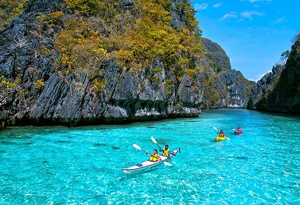 People kayaking on the clear blue waters of El Nido, surrounded by rocky cliffs and lush greenery under a bright sky.