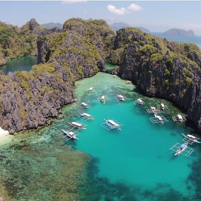 Aerial view of a turquoise lagoon in El Nido, surrounded by rocky cliffs, with several small boats on a scenic boat tour gliding across the water.