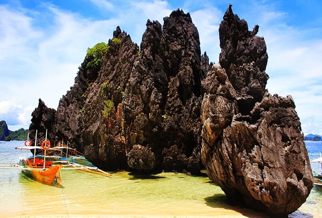 A wooden boat from an El Nido boat tour is moored near a large, jagged rock formation on a sandy shore, with clear blue skies in the background.