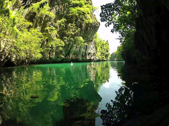 A tranquil river surrounded by lush green cliffs and trees reflects in the clear water under a bright sky, reminiscent of an El Nido boat tour.