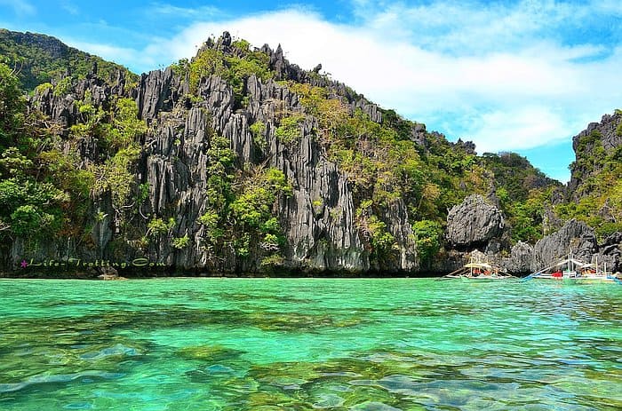 El Nido's clear turquoise waters welcome you to explore their beauty, as boats near rocky cliffs and lush green vegetation glide under a brilliant blue sky. Embark on a captivating boat tour to witness this stunning scenery firsthand.