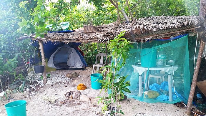 A small camping setup in a lush, green area near the starting point for island hopping adventures. A blue tent is partially covered with a palm-leaf roof, while nearby, a table with cooking utensils rests under a mosquito net. Plastic chairs and buckets are scattered around on the sandy ground.