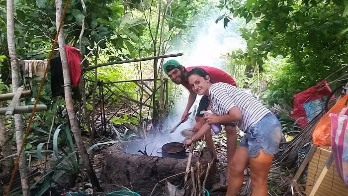 Two people are cooking over an open fire in a forested area, possibly during a break from boat tours. Smoke rises as they immerse themselves in the joys of outdoor activity, surrounded by trees and shrubs.