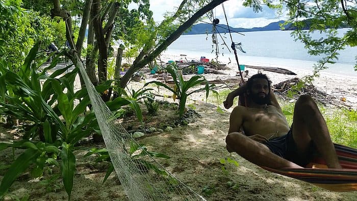 A person relaxes on a hammock tied between trees on a sandy beach, perfect for recharging before an afternoon of island hopping. Surrounding greenery and a calm sea with distant mountains enhance the serene setting. Sparse beach items and driftwood are visible on the shore.