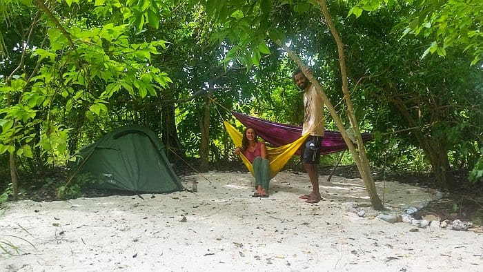 Two people are smiling at a campsite in a forest setting. One is lounging in a colorful hammock, while the other stands beside it. After an adventurous day of island hopping, they relax beside their green tent pitched on the sandy ground, surrounded by lush green trees.