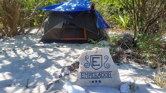 A tent is set up on a sandy beach on Dimancal Island, surrounded by trees and shrubs. In the foreground, there's a stone with "Emperador" and star symbols written on it, partially shaded by greenery—a perfect spot to relax after a day of island hopping or boat tours.