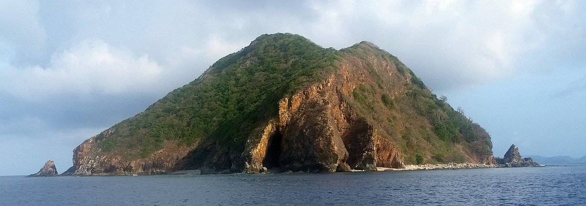A rocky island covered with lush greenery rises from the ocean near Twin Lagoons, featuring a large cave-like formation on its cliff face. The sky above is partly cloudy, casting a serene atmosphere over the tranquil water—a perfect spot for island hopping in Coron.