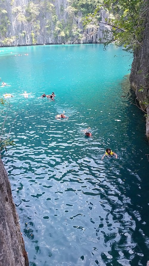Swimmers wearing life vests are enjoying the turquoise waters of Twin Lagoons, surrounded by rocky cliffs and lush greenery. Island hopping boat tours reveal this serene paradise where sunlit water reflects the vibrant blue sky, creating an inviting atmosphere.