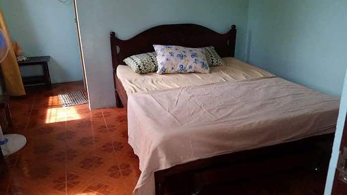 A simple bedroom with a double bed covered in beige bedding evokes a rural charm. Two pillows with floral patterns rest against the wooden headboard. The room has a tiled floor, a small side table, and light green walls. Sunlight reminiscent of Coron streams in from the left.