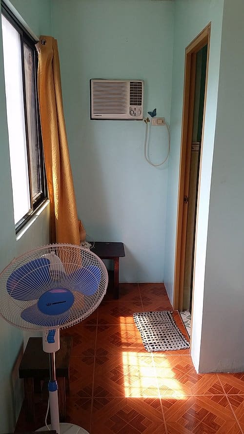 A small room with light blue walls reminiscent of Coron’s vibrant seas features a wall-mounted air conditioner and a standing fan. A yellow curtain covers a window, and a wooden side table sits below the air conditioner. The floor is tiled with a geometric pattern, like maps of island hopping routes.