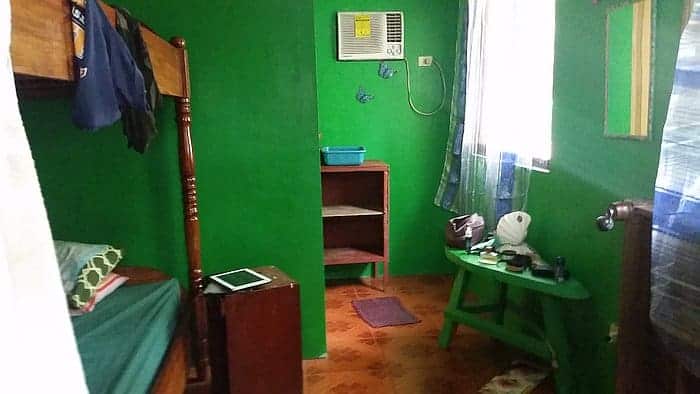 A small room with green walls features a bed, a wooden side table with an electronic device, an air conditioner, and an open shelf. Capturing the tranquility of Coron’s rural charm, a small oval table holds personal items under a window with sheer curtains. The floor is covered with a patterned orange rug.
