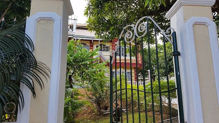 A black wrought iron gate flanked by two light-colored pillars opens to reveal a large, red and yellow house surrounded by lush green trees and plants, reminiscent of the vibrant scenery one might encounter while island hopping in Coron. The scene is bright, capturing a tranquil and inviting atmosphere.