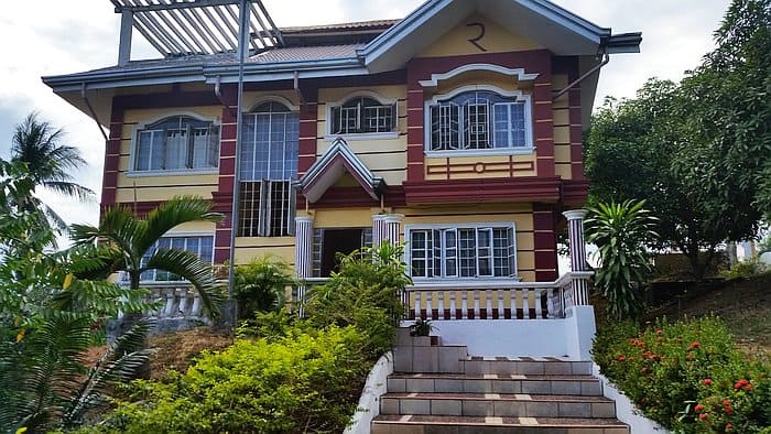 A large, two-story house with yellow and red exterior colors sits in a picturesque rural setting. It boasts multiple windows and a central entrance leading up from lush greenery, including bushes and palm trees. The sky is partly cloudy, evoking an inviting vibe reminiscent of dreamy boat tours near Coron.