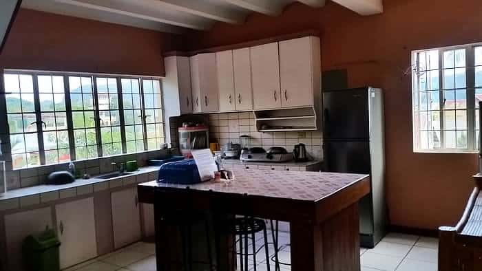 A kitchen with large windows offers a view of rural greenery, complemented by a refrigerator, white cabinets, and a tiled countertop island with stools. Various kitchen appliances and the scene outside evoke the tranquil charm of Coron, much like returning from boat tours.