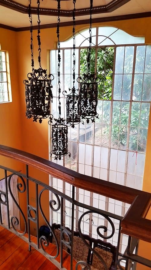 A staircase landing features ornate metal railings and a group of vintage-style hanging lanterns. Large windows allow natural light to flood the area, offering a view of greenery reminiscent of a serene rural landscape. The walls are painted in a warm, yellow tone.