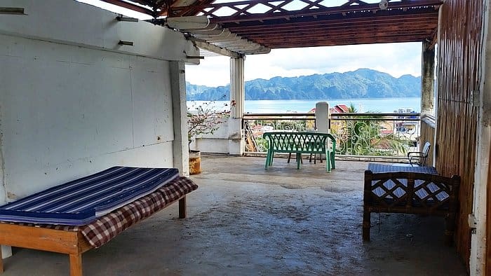 A covered outdoor patio with a wooden roof, featuring a cushioned bench and a table with chairs, offers breathtaking views of the sea and distant green mountains in Coron. Its rustic charm is enhanced by bamboo decorations on one side, making it the perfect spot to unwind after island hopping adventures.