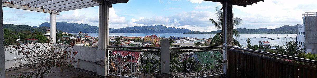 A panoramic view of a coastal town with lush green mountains and a calm blue sea in the background. The foreground features a partially covered balcony with a railing, some potted plants, and a few scattered clouds, perfect for watching boat tours glide by.