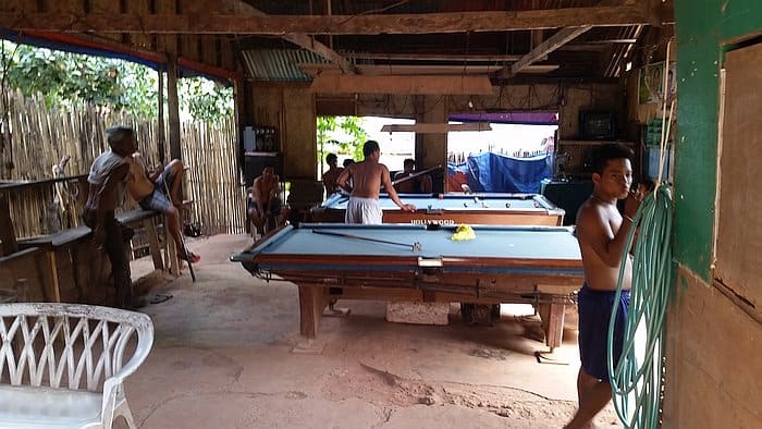 In a rural open-air area with wooden walls and a thatched roof, a group of people, some shirtless, are playing pool on two tables. A man stands near a green hose while others sit or watch, evoking the laid-back vibe of island hopping in Coron.