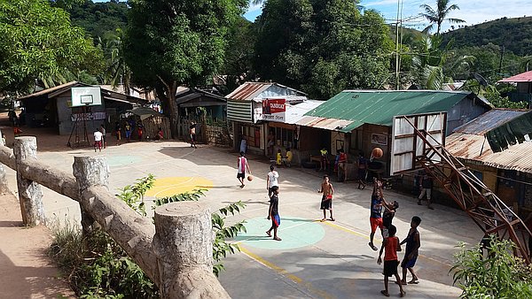 A group of people playing basketball on an outdoor court in a rural village setting of Coron. The court is surrounded by trees and rustic buildings, set against a mountainous landscape under a blue sky.