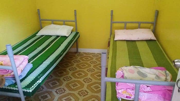 A small, rural room with yellow walls features two single beds adorned with striped green mattresses, each accompanied by a pillow and folded blankets at the foot. The floor showcases patterned tiles in shades of brown and beige, reminiscent of Coron’s earthy charm.