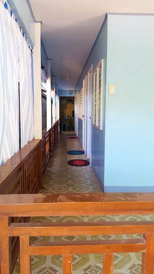 A narrow hallway with light blue walls and patterned tile flooring evokes the charm of a seaside retreat. Wooden railings line the left, and vibrant round mats enhance the doors on the right. At the end, a chair awaits like a traveler after island hopping in Coron.