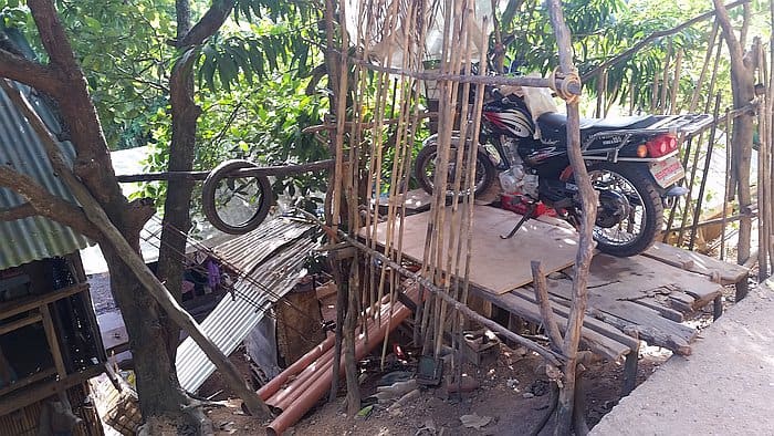 A motorcycle is precariously parked on a wooden platform supported by bamboo poles above an uneven surface. This rural structure, nestled next to trees, appears makeshift with corrugated metal below, reminiscent of the rustic charm found in Coron while island hopping.