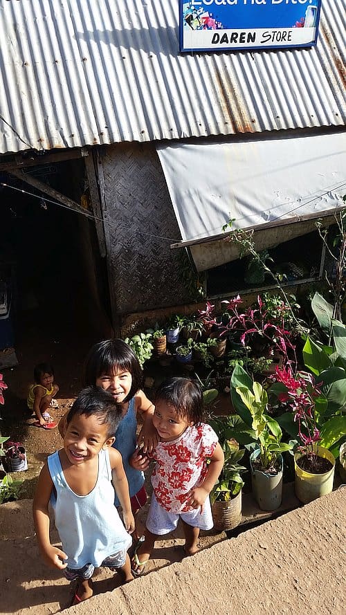 Three children smile on a dirt path surrounded by potted plants in the rural setting of Coron. Behind them stands a rustic building with a corrugated metal roof and a sign reading "Daren Store," as sunlight casts shadows on this charming scene.