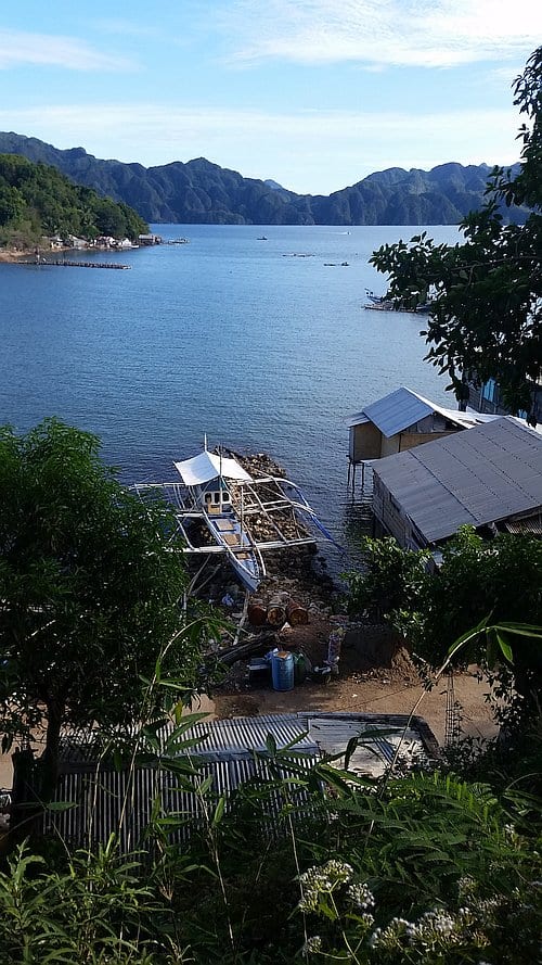 A serene coastal scene in Coron with a wooden boat docked along a small pier, surrounded by lush green foliage. Crystal-clear blue waters stretch to distant, rugged mountains under a partly cloudy sky. Coastal buildings are visible near the shore, inviting you to start your island hopping adventure.