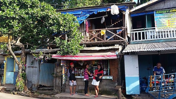 A small shop in a rustic, rural street setting with a tin roof and wooden facade. Three people stand in front, chatting and smiling. A fourth person is near a blue-painted section, typical of Coron architecture. A leafy tree adds to the laid-back charm on the left.