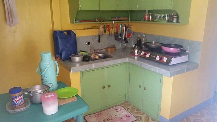 A small rural kitchen with green cabinets, a countertop adorned with cooking utensils, and a stove featuring a pink pan. There's a table with a thermos, dishes, and food containers. The walls are yellow, and a rug lies on the floor. A blue bag hangs by the sink.