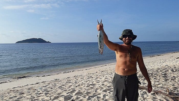 A shirtless man in sunglasses and a hat stands on a sandy beach, holding a fish in one hand. The ocean and a small island promise perfect spots for island hopping boat tours under the expansive blue sky.