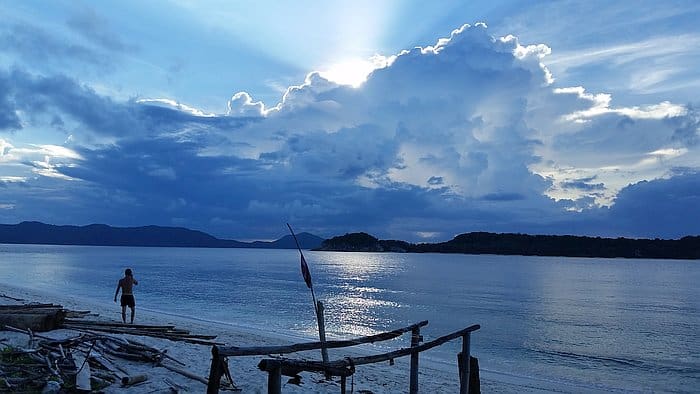 A person walks along a tranquil beach at sunset, dreaming of island hopping adventures. Dramatic clouds partially cover the sun, casting a blue hue over the sky and reflecting on the calm sea. A simple wooden structure stands in the foreground, with distant hills visible on the horizon.