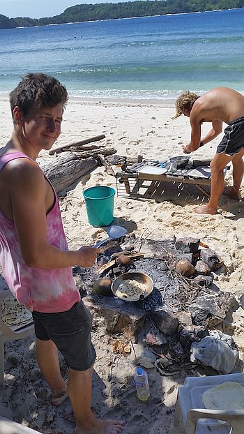 Two people are cooking on a beach, enjoying island hopping as they savor the moment. One is expertly using a pan over an open fire with various items scattered around, while the other preps food on a beach chair. The ocean and trees set a serene backdrop for this culinary adventure.