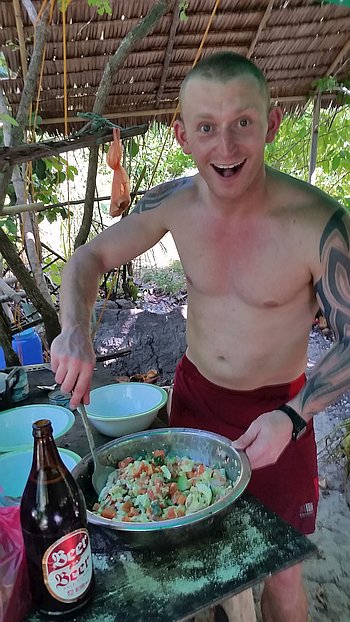 A shirtless person with a tattoo is smiling and mixing a large bowl of food outdoors, perhaps after an adventurous day of island hopping. A bottle labeled "Beer" sits on the table next to bowls, set against a lush, leafy background.