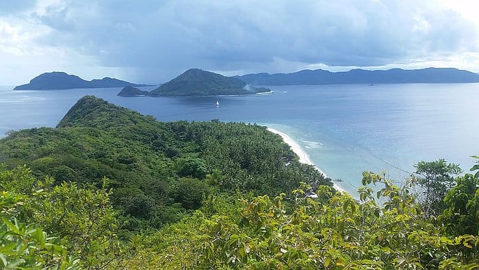 A scenic view of a lush green island with dense forests and a sandy beach surrounded by blue ocean waters invites you to explore further. Island hopping reveals several smaller islands under the cloudy sky, and nearby, boat tours drift quietly near the shoreline.