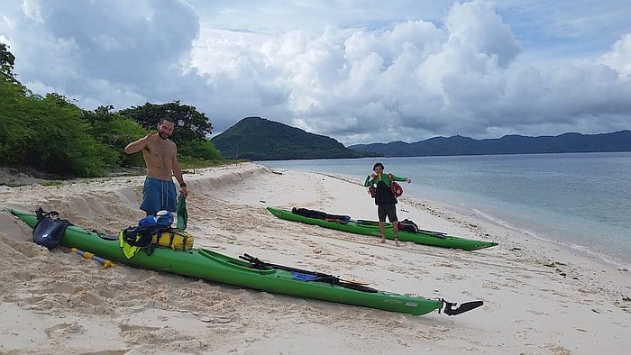 Two people stand on a sandy beach next to two green kayaks, ready for an island-hopping adventure. One gives a thumbs up while the other prepares gear. The ocean is calm with lush green hills in the background under a cloudy sky, promising an ideal day for boat tours.